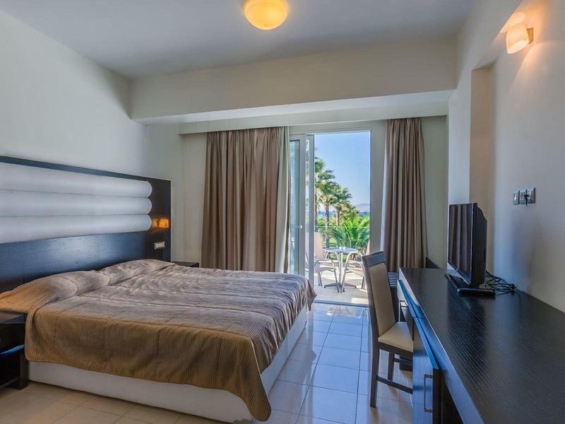 Jasny pokój hotelowy z łóżkiem podwójnym, biurkiem i balkonem z widokiem na palmy.