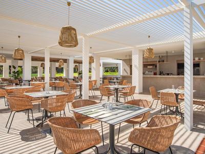 Hell gestaltetes Restaurant mit weißen Wänden und Holzmöbeln unter weißem Lamellendach.