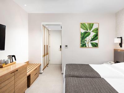 Modernes Schlafzimmer mit Doppelbett, Holzmöbeln, TV und Pflanzenbild an heller Wand.