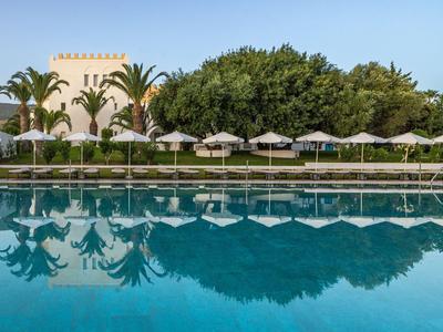 Großer Pool mit Sonnenliegen und Schirmen vor Palmen und einem Hotelgebäude am sonnigen Tag.