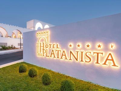 Beleuchtetes Hotel-Schild "Hotel Platanista" an weißer Wand mit grünem Rasen und blauem Himmel.