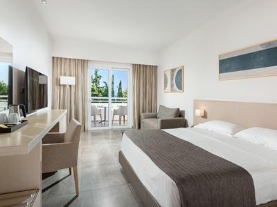 Helles Hotelzimmer mit großem Bett, Schreibtisch, Fernseher und Balkon mit Blick ins Grüne.