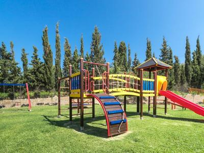 Bunter Spielplatz mit Rutsche, Klettergerüst und Schaukel auf grünem Rasen unter blauem Himmel.