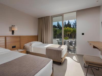 Helles Hotelzimmer mit zwei Betten, Balkon und Blick auf grüne Bäume.