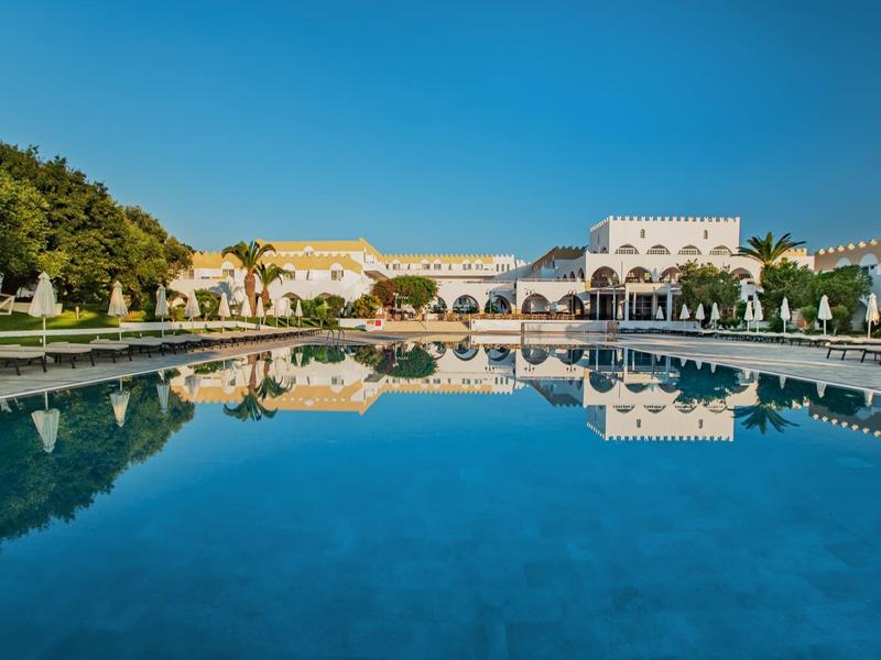 Weißes historisches Gebäude mit Bögen spiegelt sich in einem klaren, blauen Wasserbecken unter blauem Himmel.