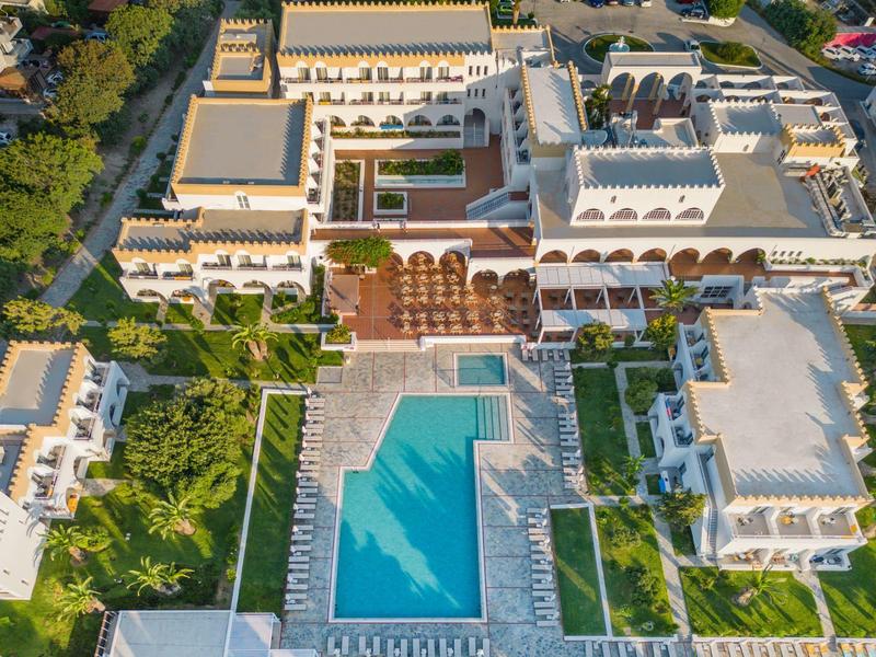 Große weiße Hotelanlage mit blauem Pool, umgeben von Palmen und Liegestühlen, aus der Vogelperspektive.