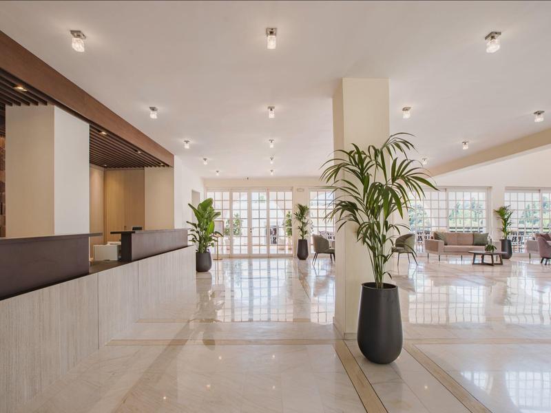 Große helle Hotellobby mit Marmorboden, Empfangstresen links und grünen Pflanzen in Vasen.