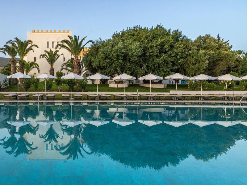 Ein ruhiger Pool mit blauer Wasseroberfläche, Sonnenschirmen und Palmen vor einem Hotel.