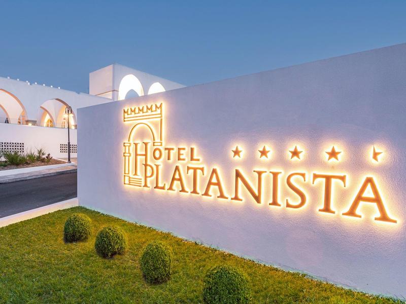 Beleuchtetes Hotel-Schild "Hotel Platanista" an weißer Wand mit grünem Rasen und blauem Himmel.