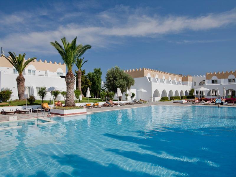 Großer blauer Pool vor weißen Gebäuden mit Palmen und Sonnenschirmen unter blauem Himmel.