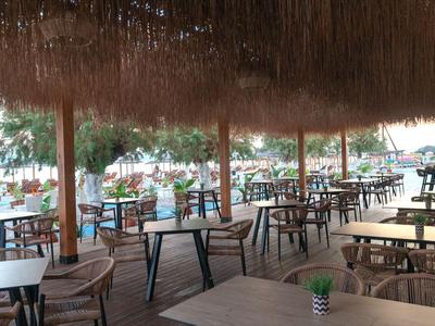 Überdachte Terrasse mit Stroh- und Holzstuhlgruppen, Tischen und Blick auf Palmen und Wasser.