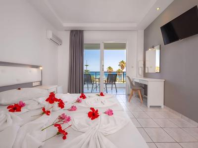 Helles Hotelzimmer mit weißem Bett, roten Blumen, Balkon und Meerblick.