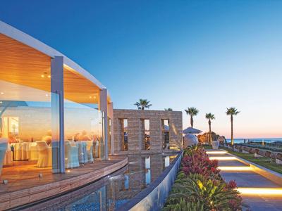 Luxuriöse Terrasse mit Glaswänden, Pool, Palmen und Meerblick bei klarem Himmel am Abend.