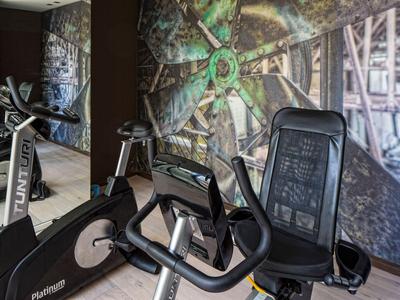 Sala fitness con due cyclette davanti a una parete con grande motivo industriale.