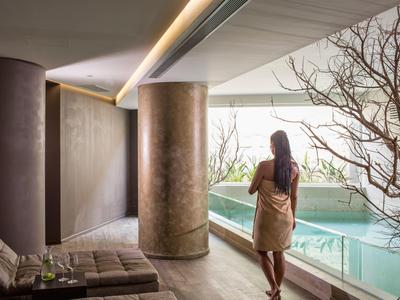 Žena v ručníku stojí u vnitřního bazénu s velkými sloupy a minimalistickou výzdobou.