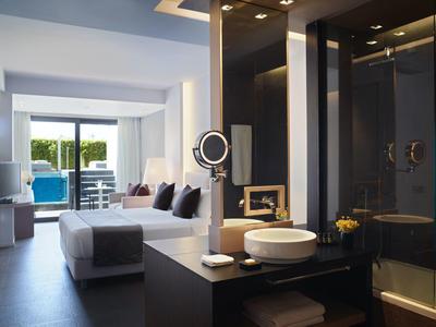 Moderní hotelový pokoj s postelí, umyvadlem, posezením a výhledem na terasu s bazénem.