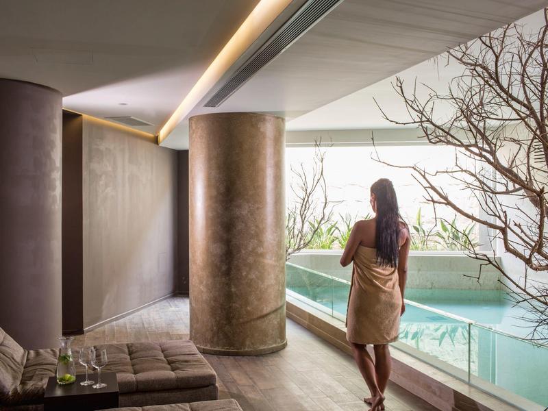 Žena v ručníku stojí u vnitřního bazénu s velkými sloupy a minimalistickou výzdobou.