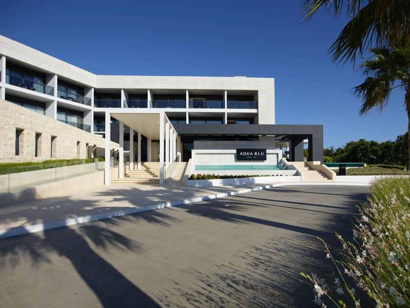 Moderní hotel s bazénem, bílým a černým fasádním povrchem, obklopený zelení pod jasně modrou oblohou.