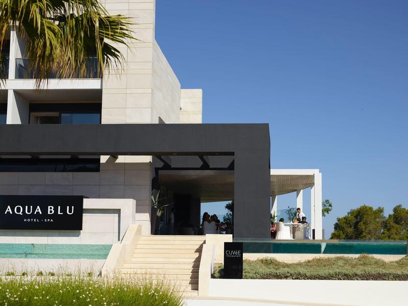 Vstup do hotelu s nápisem Aqua Blu, moderní architektura, schody a palma pod modrou oblohou.