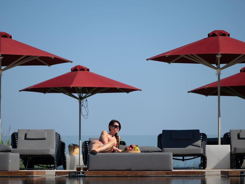 Osoba odpočívá na lehátku pod červenými slunečníky u bazénu za jasného dne.