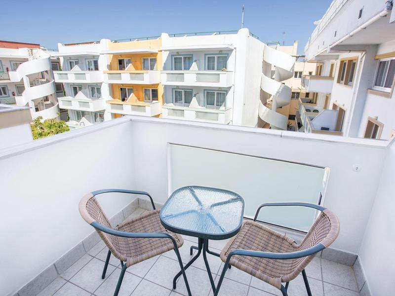 Balcón con dos sillas de mimbre y mesa pequeña de vidrio, edificio moderno blanco al fondo.