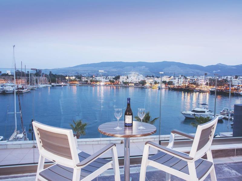 Vista de un puerto con veleros desde una terraza con mesa y dos sillas al atardecer.