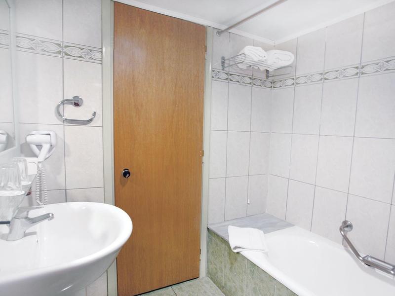 Pequeño baño con bañera, lavabo, puerta de madera y azulejos blancos en las paredes.