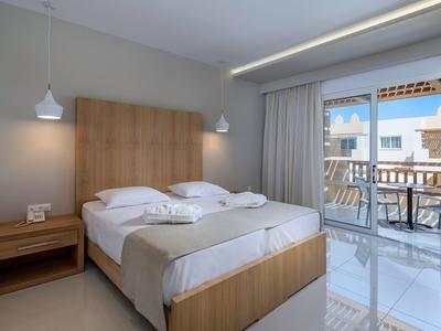 Habitación de hotel luminosa con cama doble, muebles de madera y balcón con vista a edificios blancos.