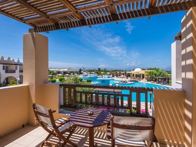 Balkon mit Holzmöbeln blickt auf großen Pool und Hotelgebäude unter blauem Himmel.