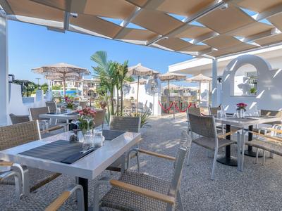 Freiluftrestaurant mit Tischen, Stühlen und Sonnenschutz, umgeben von Palmen und Gebäuden.