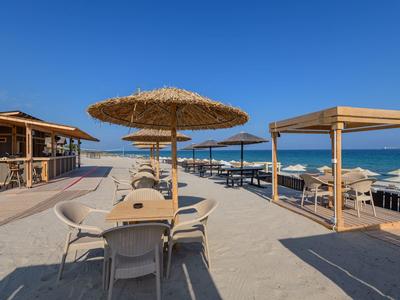 Terraza de hotel frente a la playa con sombrillas de paja, sillas y mesas bajo un cielo azul claro.