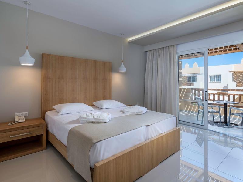 Habitación de hotel luminosa con cama doble, muebles de madera y balcón con vista a edificios blancos.