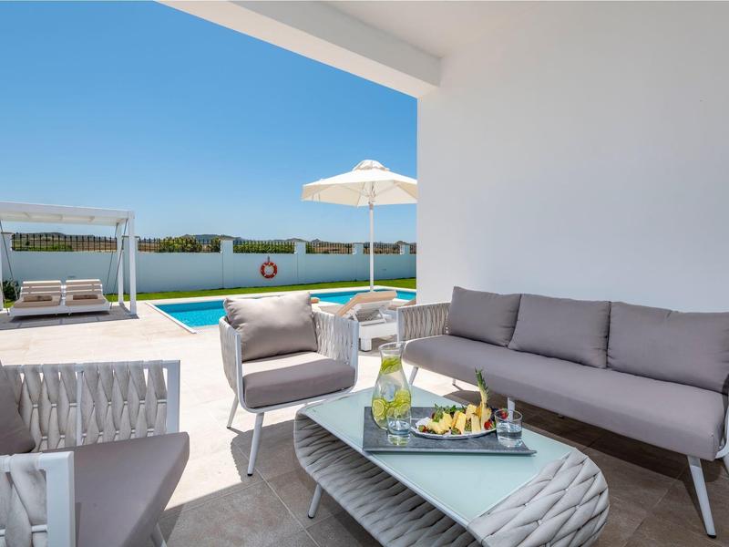 Elegante zona de estar exterior con sofás grises, mesa y vista a la piscina y cielo azul.