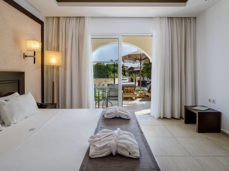 Habitación de hotel luminosa con cama doble, batas en la cama y vista al patio con sillas y sombrilla.