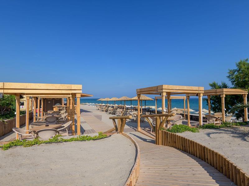 Pasarelas y pérgolas de madera conducen a una playa de arena con vista al mar azul bajo cielo despejado.