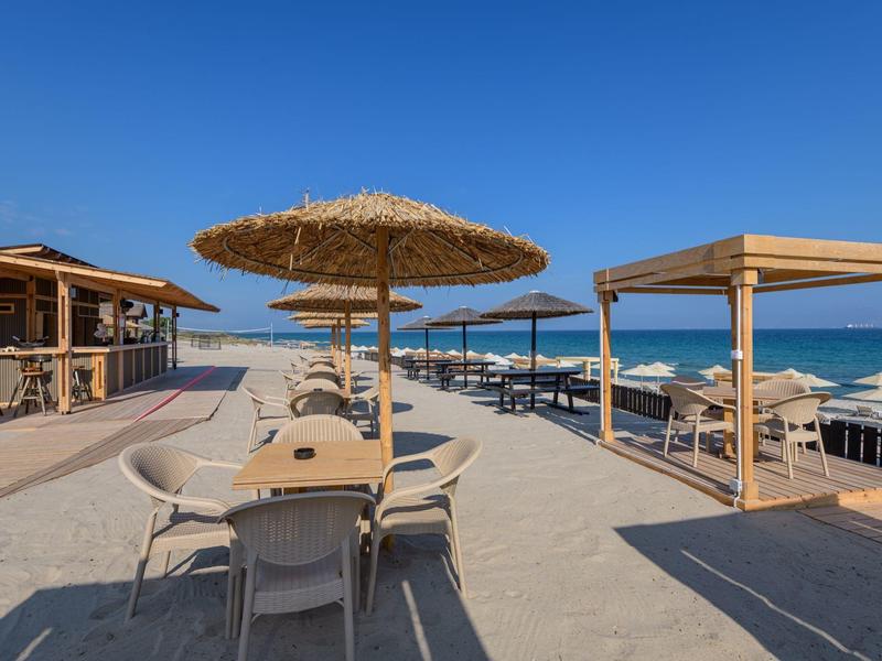 Terraza de hotel frente a la playa con sombrillas de paja, sillas y mesas bajo un cielo azul claro.