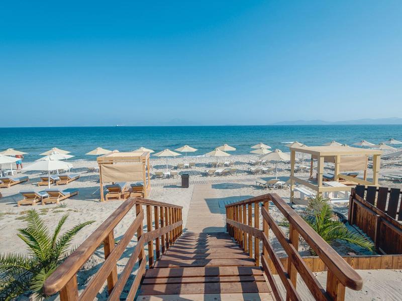Playa con sombrillas, tumbonas y pasarela de madera hacia el mar en un día despejado.