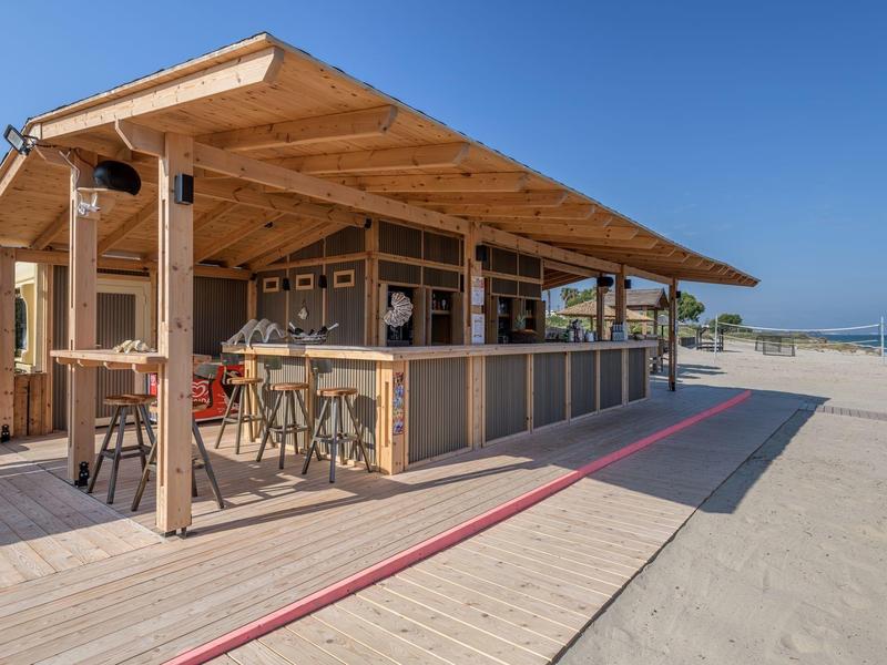 Bar de playa con estructura de madera, taburetes y techo, rodeada de arena bajo un cielo despejado.