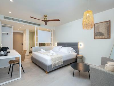 Helles Hotelzimmer mit großem Bett, sofa, runden Tischen, Schreibtisch und warmem Deckenlicht.
