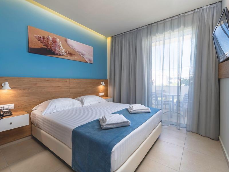 Jasný hotelový pokoj s manželskou postelí, modrou zdí a balkonem se záclonami.