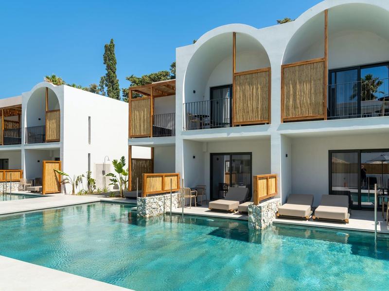 Moderní hotel s bílou budovou a bazénem, obklopený lehátky a zelenými rostlinami.