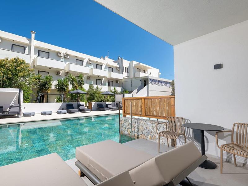 Pohled z terasy s lehátky na bazén a bílou hotelovou budovu pod modrou oblohou.