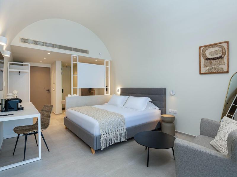 Světý hotelový pokoj s manželskou postelí, psacím stolem, křeslem a moderní koupelnou.