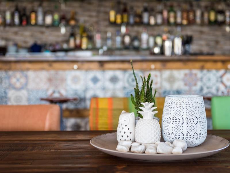 Talíř s bílými keramickými dekoracemi a ananasem na dřevěném stole v kavárně s barem v pozadí.