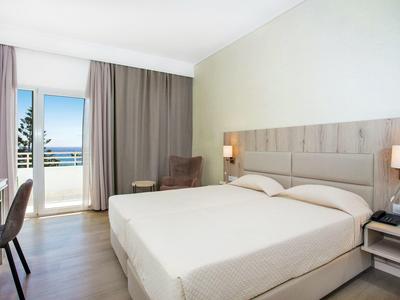 Helles Hotelzimmer mit großem Doppelbett, braunem Vorhang, Stuhl und Balkon mit Meerblick.