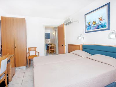 Helles Hotelzimmer mit zwei Betten, Schrank, Stühlen, Schreibtisch und farbenfrohem Kinderbild an der Wand.