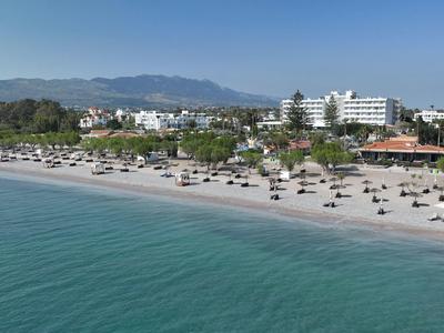 Strand mit Sonnenschirmen und Liegen vor grünen Bäumen und Gebäuden unter blauem Himmel.