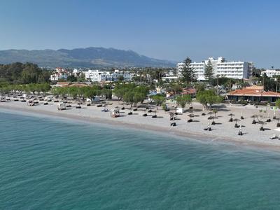 Strand mit Liegestühlen und Sonnenschirmen vor einem Hotel, ruhiges blaues Meer im Vordergrund.