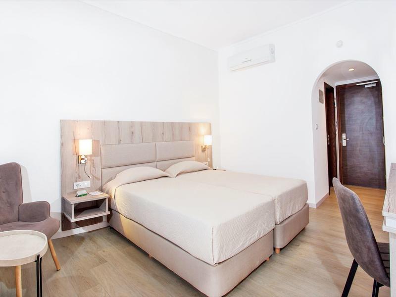 Světý hotelový pokoj s manželskou postelí, dvěma křesly, malým stolem a televizí na zdi.