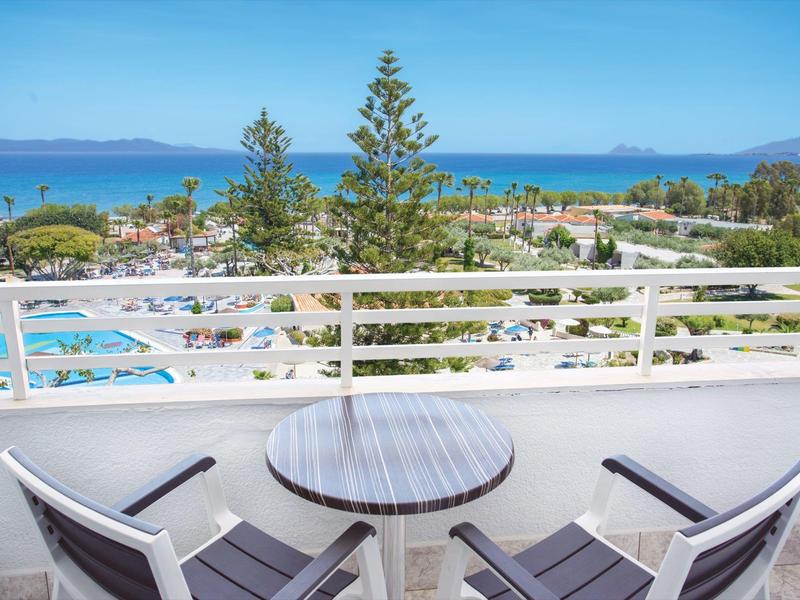 Výhled z balkonu se stolem a židlemi na bazén, stromy a moře v pozadí.